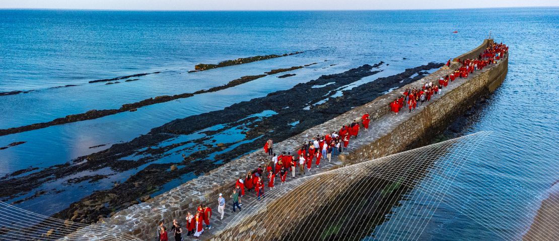 Students in red gowns walking on an old stone pier, surrounded by blue sea.